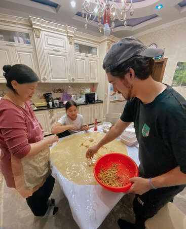 Three people are preparing food together in a cozy kitchen. They are spreading vegetable filling onto a large sheet of dough, smiling and enjoying the cooking process.