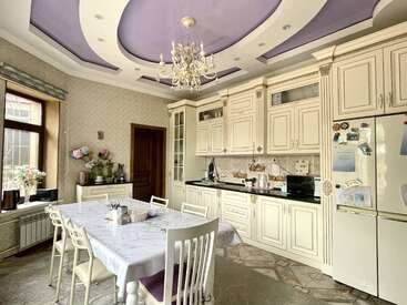This elegant kitchen features cream cabinets, a lavender coffered ceiling, chandelier, marble-topped dining table, white chairs, large windows, and a fridge adorned with magnets.