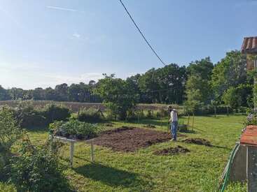 Ein Mann steht in einem sonnigen Garten und bereitet ein Stück Erde für die Bepflanzung vor. Daneben steht ein Tisch mit Topfpflanzen, umgeben von üppigem Grün und Feldern.