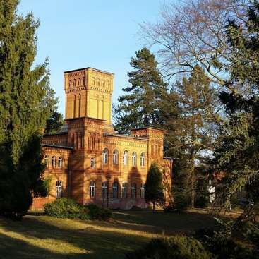 The image depicts a majestic yellow brick building with a tower, surrounded by trees and a lush lawn, set against a clear blue sky.