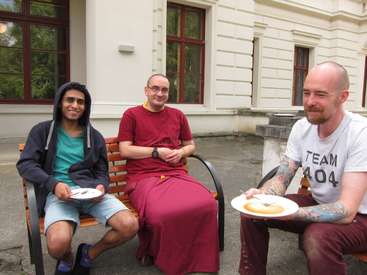 The image depicts three men sitting on a bench, each holding a plate of food, in front of a white building with windows and a door.