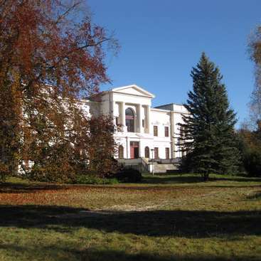 The image depicts a large white building with columns, situated behind a grassy area and trees, set against a clear blue sky.