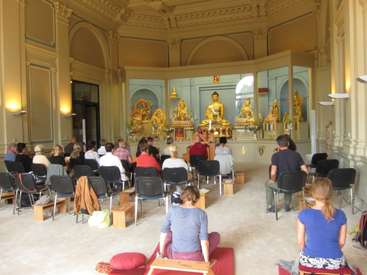 The image depicts a serene room with a group of people seated in chairs, facing a large golden Buddha statue, surrounded by smaller statues and a peaceful atmosphere.