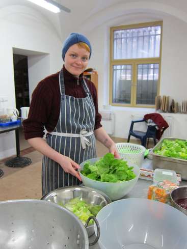 The image depicts a woman in a kitchen, wearing a blue beanie and apron, standing behind a counter with large bowls of lettuce, preparing a meal.