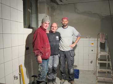 Three men stand in a bathroom under construction, surrounded by white tiles and tools, posing for a photo with their arms around each other\'s shoulders.