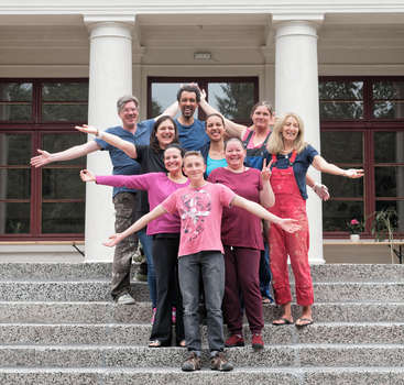 The image depicts a group of nine people standing on the steps of a building with white columns, posing for a photo with their arms outstretched.