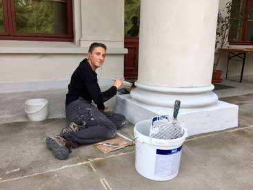A man is sitting on the ground, painting a white column with a paintbrush, wearing a black shirt and grey pants, with a bucket of paint nearby.