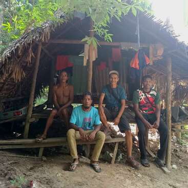 L'image représente quatre hommes assis sur un banc en bois devant une hutte, entourée d'une végétation luxuriante et d'arbres. Le toit de chaume de la hutte est visible.