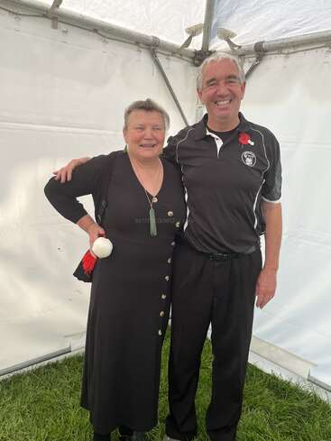 A smiling woman and man stand together inside a tent. The woman wears a black dress and green pendant, the man wears a black sports shirt with a poppy.