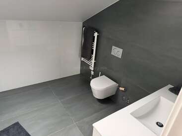 Das ist ein modernes Badezimmer mit grauen Fliesen, einem Wand-WC, einem weißen Waschbecken, einem Handtuchhalter mit einem dunklen Handtuch und einem minimalistischen, klaren Design.