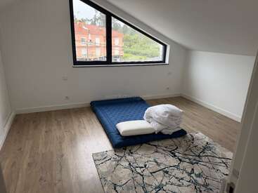 Ein minimalistisch eingerichtetes Schlafzimmer im Dachgeschoss mit einem großen Schrägefenster, einer blauen Matratze, ordentlich gefalteter Bettwäsche, einem Kissen und einem modernen, abstrakten Teppich auf Holzböden vor weißen Wänden.