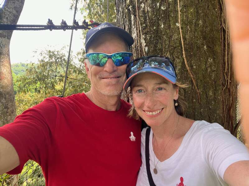 Ein lächelnder Mann und eine lächelnde Frau posieren draußen für ein Selfie; sie stehen dicht beieinander unter einem Baum und tragen beide Freizeitkleidung, Hüte und Sonnenbrillen.