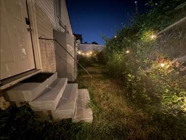 Un acogedor patio trasero iluminado por guirnaldas de luces; unos frondosos arbustos verdes bordean el camino, y unos escalones de hormigón conducen a una puerta. El cielo del atardecer es de color azul oscuro.