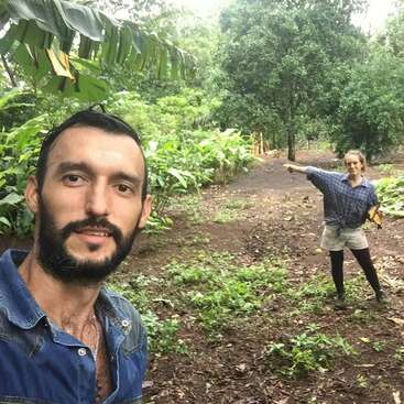L'image représente deux personnes debout dans une clairière luxuriante, l'une pointant vers la droite et l'autre prenant un selfie au premier plan.