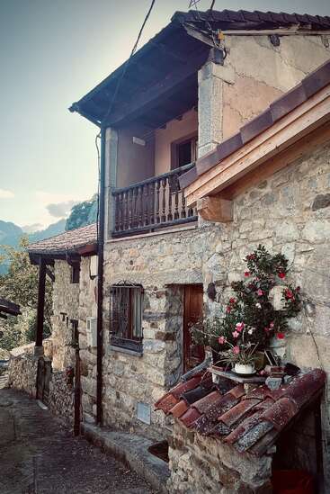 Une maison rustique en pierre avec un balcon en bois, un toit en tuiles rouges et des fleurs en pot. Nichée dans un village de montagne, entourée de verdure, elle dégage un charme d'antan.
