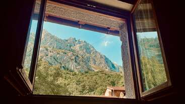 Par une fenêtre ouverte, des montagnes escarpées et des arbres verdoyants se dessinent sous un ciel bleu limpide. Un paysage paisible et ensoleillé offre une vue pittoresque et tranquille depuis l'intérieur.