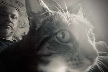 Una foto en blanco y negro muestra a un hombre sentado en un sofá con un curioso gato en primer plano, que mira fijamente al objetivo de la cámara.