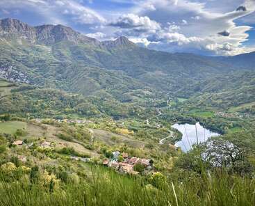 Un pintoresco valle con exuberantes colinas verdes, casas dispersas, un río serpenteante, un lago reflectante, montañas majestuosas, nubes espectaculares y una vegetación vibrante crean un paisaje tranquilo y pintoresco.