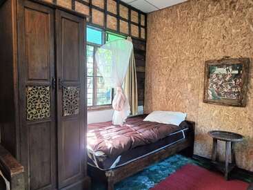 Este quarto rústico aconchegante tem uma cama de solteiro, guarda-roupa de madeira, pequena mesa redonda, paredes de madeira texturizada, janela com luz solar, cortinas brancas, porta-retratos e tapete vermelho.