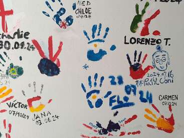 Uma parede coberta com impressões de mãos coloridas, datas e nomes como Charlie, Chloe, Lorenzo e Carmen. Alguns rabiscos e textos em vários idiomas também estão presentes, criando uma cena divertida.