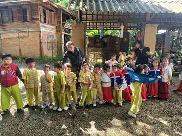 Um grupo de crianças pequenas e dois adultos estão do lado de fora, em frente a um prédio, vestidos com uniformes coloridos, parecendo estar em uma excursão escolar.
