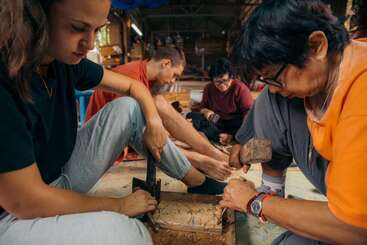 Quatro pessoas sentam-se juntas no chão, concentradas na escultura em madeira. Elas trabalham juntas, usando várias ferramentas, criando desenhos complexos em um ambiente de oficina acolhedor e comunitário.