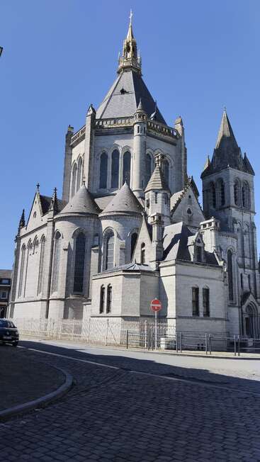 A grand stone cathedral with intricate gothic architecture, pointed towers, arched windows, and a tall spire topped with gold, stands majestically under a clear blue sky.