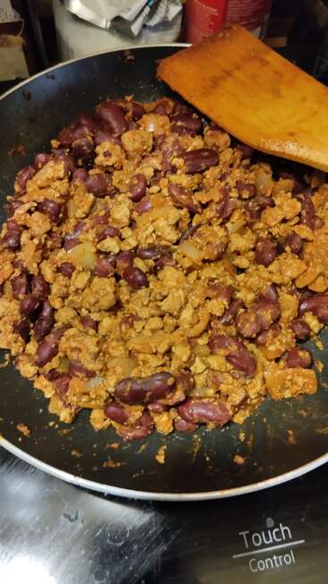 A frying pan filled with cooked kidney beans, ground meat, onions, and spices. A wooden spatula rests on the pan. Stove's touch control panel visible.