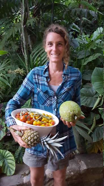 Eine Frau steht vor üppigem Grün, hält eine Schüssel mit Tomaten, eine Ananas und einen Kürbis in der Hand und hat ein warmes Lächeln im Gesicht.