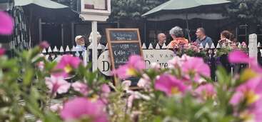 Bright pink flowers fill the foreground, while people sit and converse at an outdoor café. A chalkboard menu stands by white picket fencing under umbrellas.