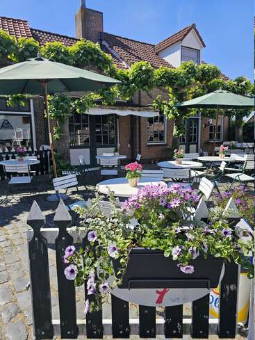 A charming outdoor café with white tables, green umbrellas, and blooming flowers. Cozy brick house in the background, vines climbing the facade, inviting atmosphere, sunny day.