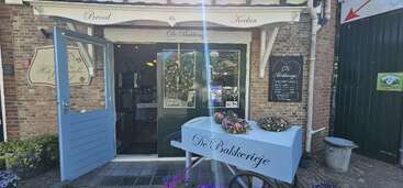 This image shows the entrance of a rustic bakery called "De Bakkerijeje" with flowers, a blue cart, and inviting open doors surrounded by brick walls.