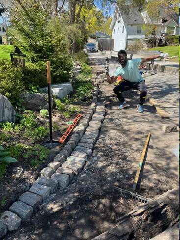 Un homme pose joyeusement tout en travaillant sur un chemin de jardin bordé de briques en pierre. Des outils et un niveau se trouvent à proximité, et la végétation entoure la zone de travail.