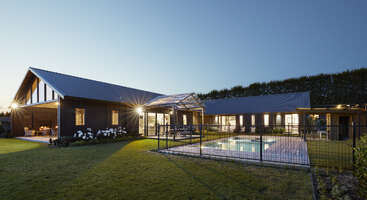 The image depicts a modern, single-story house with a dark exterior and a large yard, featuring a pool and patio area, set against a serene blue sky.