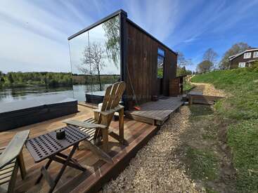 Eine gemütliche Holzhütte mit Glaswänden steht am Ufer eines ruhigen Sees. Auf der Terrasse stehen zwei Holzstühle und ein kleiner Tisch, umgeben von üppigem Grün.