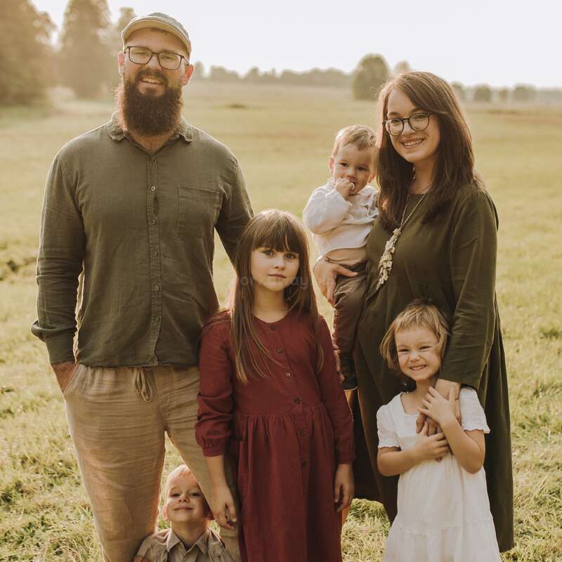 A smiling family of five stands together outdoors in a sunny field. The parents and three children are posing warmly, dressed in earthy tones, enjoying nature.