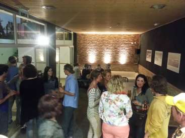 A imagem mostra uma reunião de pessoas em uma sala com uma parede de tijolos, com um sofá e obras de arte nas paredes, com algumas pessoas segurando bebidas.