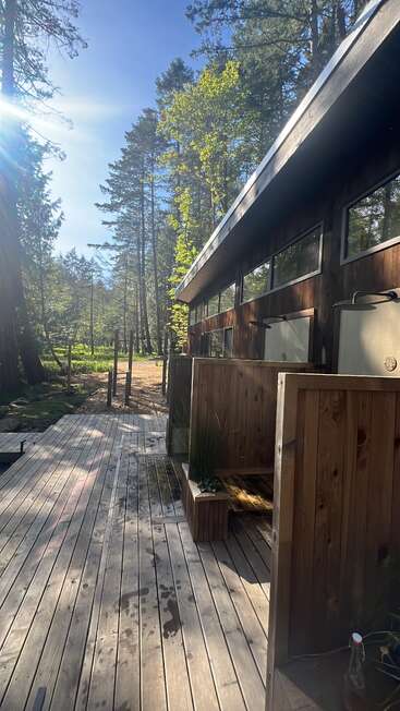 La luz del sol se filtra a través de los altos árboles del bosque y baña una terraza de madera. Junto a una cabaña moderna hay una ducha exterior revestida de madera, que transmite tranquilidad y cercanía a la naturaleza.