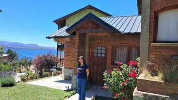Une femme se tient devant une charmante maison en briques au toit métallique, entourée de jardins luxuriants et de fleurs rouges épanouies, surplombant un lac paisible et des montagnes.