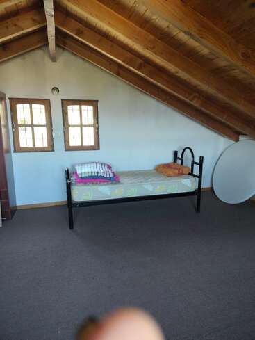 Cette chambre mansardée confortable dispose d'un plafond en bois incliné, de deux petites fenêtres, d'un lit simple avec une literie colorée, d'un sol recouvert de moquette et d'un objet rond à proximité.