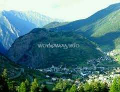 La imagen muestra una pequeña ciudad enclavada en un valle, rodeada de montañas y árboles, con una sutil marca de agua que reza "workaway.info" en el centro.
