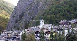 La imagen representa una pequeña ciudad enclavada en las montañas, con un prominente campanario blanco y un grupo de edificios rodeados de árboles y un acantilado rocoso.