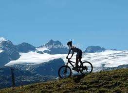La imagen muestra a un ciclista de montaña sobre una colina cubierta de hierba con las montañas nevadas de fondo en un día soleado.