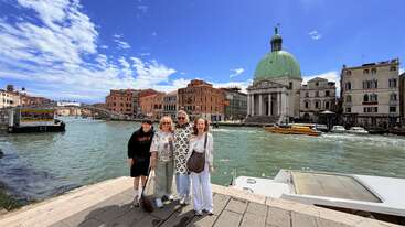 Four people pose by a canal in Venice, Italy. Historic buildings, including a church with a green dome, and boats are visible under a bright, partly cloudy sky.