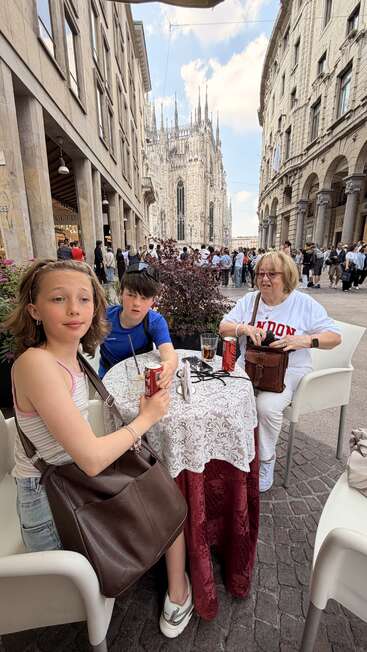 Three people sit at an outdoor café table, drinking sodas. Busy street and iconic cathedral in the background, evoking a lively European city atmosphere.