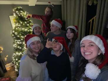 Six people wearing Santa hats smile and pose in front of a lit Christmas tree. One person stands decorating the tree. The room feels joyful and festive.