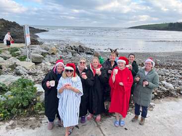 A group of women in festive hats and robes stand on a rocky beach, smiling and holding mugs, with the sea and cloudy sky behind them.
