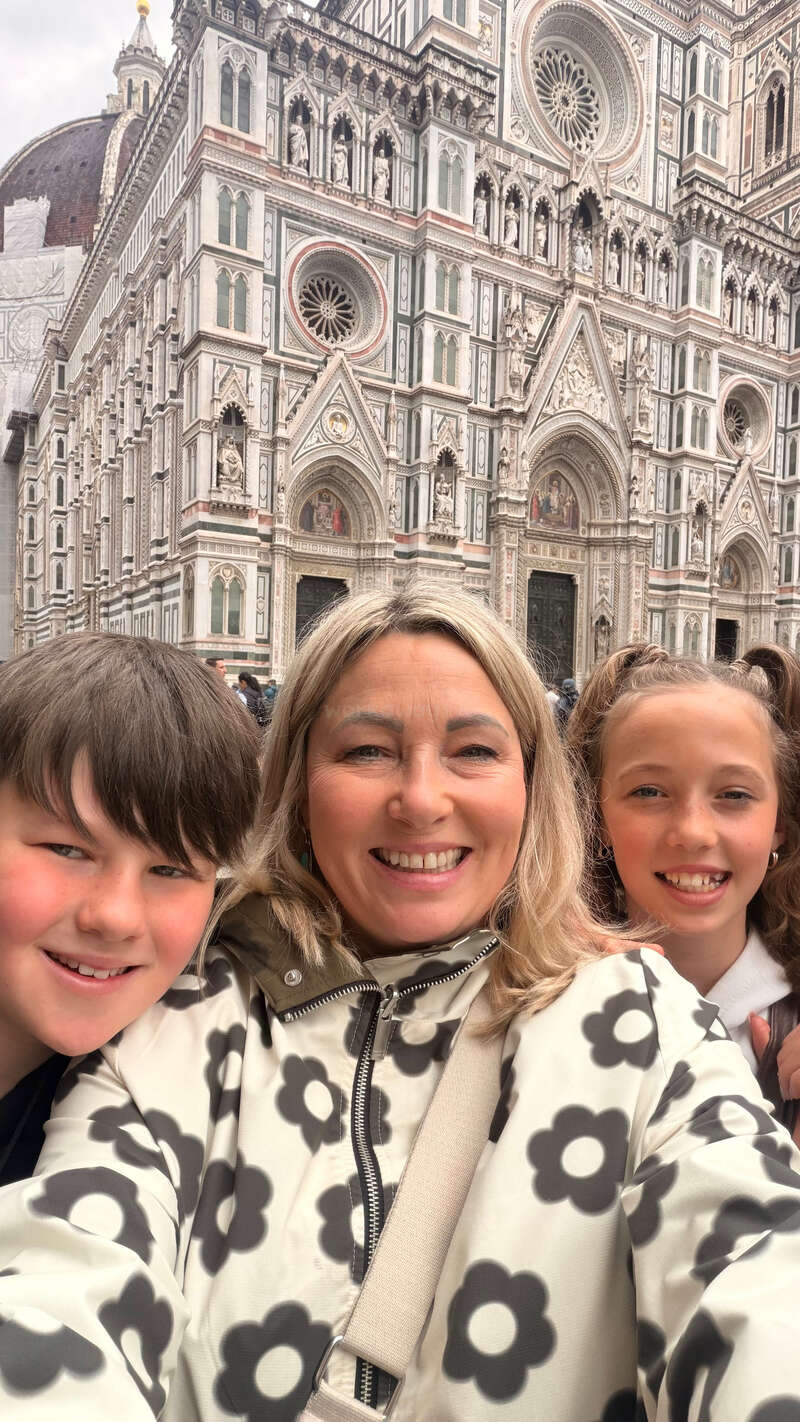 A smiling woman and two children take a selfie in front of the ornate Florence Cathedral, featuring stunning Gothic architecture, rose windows, and intricate decorative details.