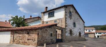 This image shows a rustic stone house with a red-tiled roof, surrounded by other traditional buildings, under a blue sky with scattered clouds. Peaceful location.
