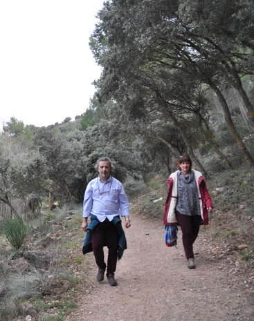 The image depicts two individuals strolling along a path, with a body of water visible in the background, surrounded by lush greenery and trees. The scene exudes a sense of serenity and tranquility.